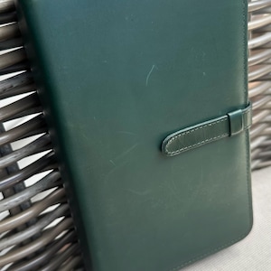 May include: A dark forest green rectangular notebook cover with a smooth finish and visible surface scratches. The cover features a matching green leather strap closure that secures horizontally across the front panel with visible stitching.