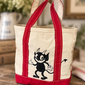 May include: A cream-colored canvas tote bag featuring two red fabric handles and a red base. The front of the bag displays a black graphic illustration of a cartoon devil character with horns, a wide grin, and a pointed tail, rendered in a vintage style.
