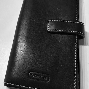 May include: Black leather wallet with white stitching and a snap closure. The wallet features the embossed "COACH" logo. The design is a classic, minimalist style, suitable for carrying cards and cash.