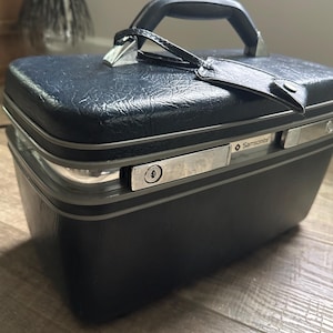 Puede incluir: Un neceser Samsonite azul marino vintage con asa negra y herrajes plateados. La maleta tiene un exterior texturizado, una cerradura y una etiqueta de equipaje de cuero. El logotipo de Samsonite es visible.