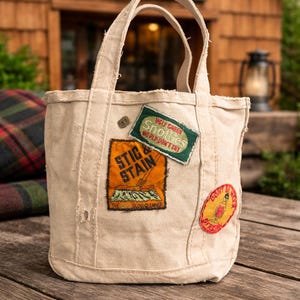 Może przedstawiać: Beżowa płócienna torba z wszytymi uchwytami i kilkoma haftowanymi naszywkami. Naszywki zawierają tekst i ilustracje, takie jak "Stic & Stain" i "Ugly Green Snooties". Torba stoi na drewnianej powierzchni.