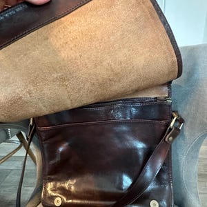 May include: A dark brown leather messenger bag with a flap closure. The bag has a shoulder strap and a front pocket. The interior is a lighter tan color. The bag is open, revealing the inside.