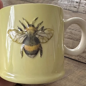 Op de afbeelding: Een gele keramische mok met een bijendesign. De bij is zwart en geel met bruine vleugels.