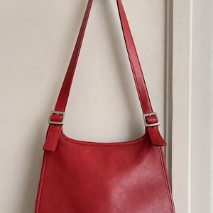 May include: A solid red leather shoulder bag with a smooth finish, featuring a long adjustable strap attached with silver-toned metal buckles on each side. The bag has a rounded bottom silhouette and hangs from a dark metal wall hook.