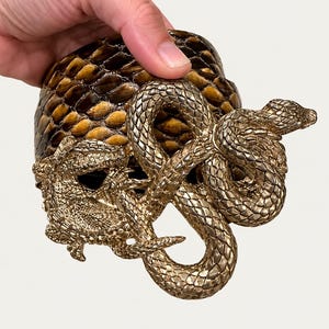 Puede incluir: Un recipiente cilíndrico con un exterior de patrón de piel de serpiente marrón y negro. La superficie está adornada con una escultura en relieve de tono dorado metálico que representa una serpiente enroscada y un lagarto con escamas detalladas.