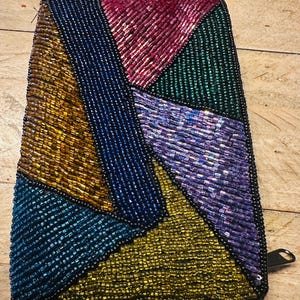 May include: A beaded clutch with a geometric design. The clutch features a patchwork of colorful beads in shades of gold, teal, blue, purple, green, and pink. The beads are sewn onto a black backing, and the clutch has a black zipper.