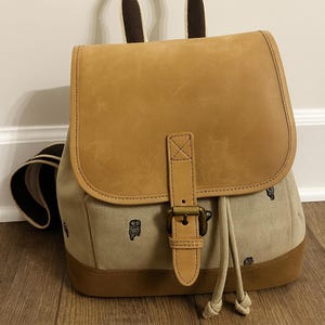May include: Tan and brown backpack with a leather flap and handle. The body of the backpack is a light tan canvas with a repeating black pattern. The bottom and handle accents are brown.