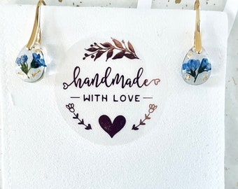 Puede incluir: Un par de pendientes de oro con un colgante de resina en forma de lágrima con flores azules prensadas y escamas doradas. Los pendientes se muestran sobre un fondo blanco con una pegatina que dice "hecho a mano con amor".
