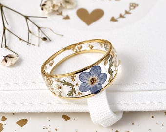 May include: A clear resin ring featuring a gold metal band edge, embedded with a single blue forget-me-not flower, green foliage, and small gold flakes. The ring is circular with a smooth, transparent finish showcasing the botanical details.
