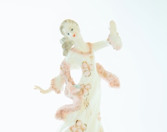 Op de afbeelding: Een porseleinen beeldje van een vrouw in een witte jurk met roze en gouden accenten. Ze draagt een sjaal met een roze en witte rand. Het beeldje staat op een witte voet.