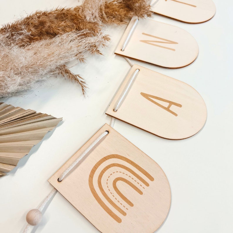 May include: A wooden banner with the letters "I", "N", "A", and a rainbow design. The banner is strung with white twine and has a wooden bead at the end.