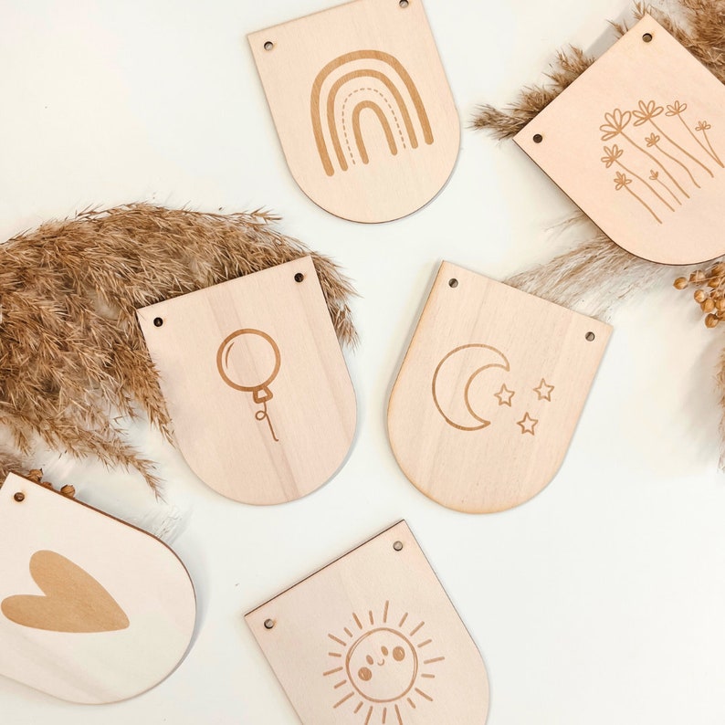 May include: A set of six wooden banner flags with various designs, including a rainbow, flowers, a moon and stars, a balloon, a heart, and a sun. The flags are all made of natural wood and have a rustic, minimalist aesthetic.