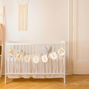 May include: White crib with a wooden name banner that reads "ANTON" hanging above it. The banner is made of individual wooden pieces with a rainbow design and gold lettering. A gray and white heart-shaped pillow is inside the crib.