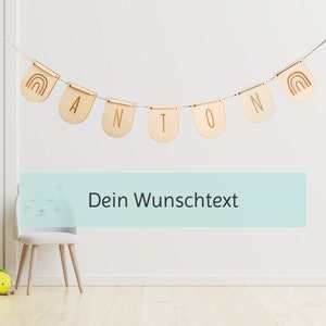 May include: Wooden banner with the name Anton and rainbow designs on each end. The banner is hanging on a white wall. The text "Dein Wunschtext" is on a light blue background below the banner.