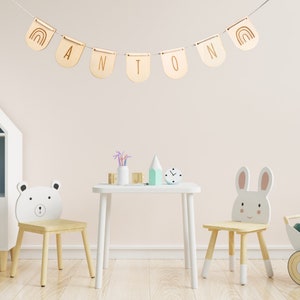 May include: A wooden name banner with the name "ANTON" spelled out in individual wooden flags. Each flag has a rainbow design on one side and a letter on the other. The banner is hanging from a white string against a light pink wall. Two wooden chairs with animal faces are in the foreground, one with a bear face and the other with a bunny face. A white table with a clock and other decorative items is between the chairs.