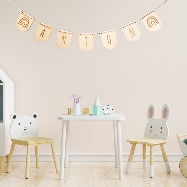 May include: A wooden name banner with the name "ANTON" spelled out in individual wooden flags. Each flag has a rainbow design on one side and a letter on the other. The banner is hanging from a white string against a light pink wall. Two wooden chairs with animal faces are in the foreground, one with a bear face and the other with a bunny face. A white table with a clock and other decorative items is between the chairs.