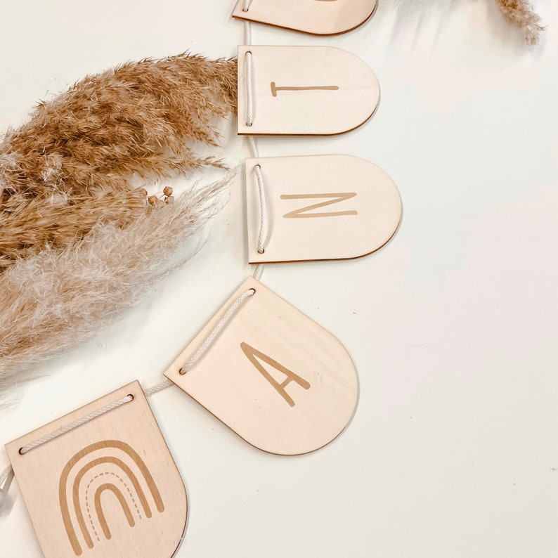 May include: A wooden banner with the word "RAIN" spelled out in cursive letters. The banner is strung together with white twine and features a rainbow design on the leftmost flag.