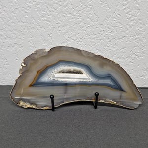 May include: A polished agate slice featuring concentric bands of gray, white, and translucent tan colors with a central crystalline geode cavity. The mineral specimen is displayed on a black metal stand with two prongs supporting the base.