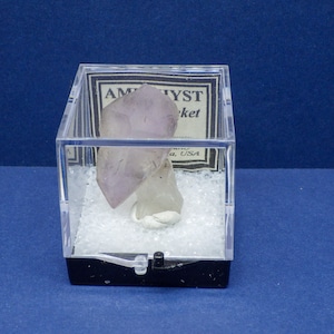 May include: A clear plastic cube with a black base displays a purple amethyst crystal on a white base. The label on the cube reads "Amethyst Pocket, Brazil, USA."