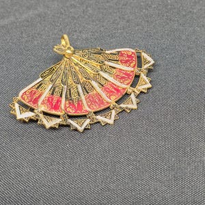 May include: A gold-toned pendant shaped like a folding fan, featuring intricate black and red enamel detailing. The fan has a scalloped edge with small triangular accents in white and gold, and a small loop at the top for attachment to a chain.