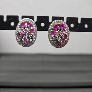 May include: A pair of oval-shaped stud earrings featuring a central cluster of vibrant pink teardrop-shaped stones and small dark blue accents, all surrounded by a border of clear, sparkling pavé-set stones in a silver-toned metal setting.