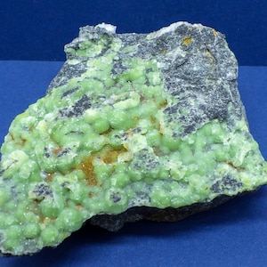 May include: A close-up of a green mineral specimen with a rough, dark gray matrix. The mineral has a botryoidal texture, with many small, rounded growths.