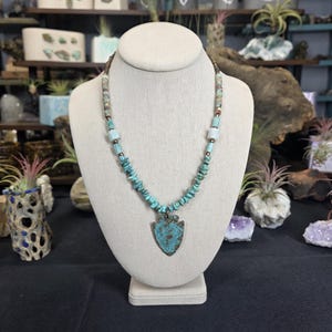 May include: Turquoise and copper-toned necklace displayed on a mannequin. The necklace features a pendant shaped like an arrowhead. The beads are a mix of turquoise and other earth-toned stones. The background shows shelves with various decorative items.