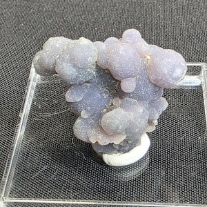 May include: A cluster of grape agate, a mineral formation, in shades of lavender and purple. The agate has a bubbly, botryoidal structure, resembling a bunch of grapes. It is displayed on a clear acrylic stand with a white base, set against a dark background.
