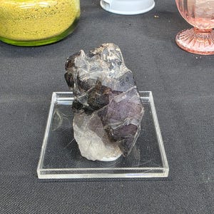 May include: A dark, multi-faceted crystal cluster with shades of purple, gray, and black, displayed on a clear acrylic square. The crystal has a rough, natural texture. The background includes a glass jar, a white pitcher, and a pink glass.