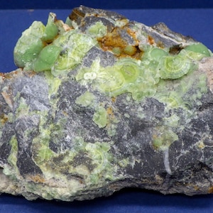 May include: A close-up of a rock with green and yellow crystals. The crystals are clustered together and have a shiny, translucent appearance. The rock is dark gray and has a rough texture.