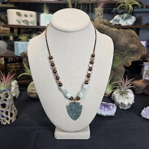 May include: A necklace with a turquoise arrowhead pendant. The necklace features a dark brown cord with alternating brown and light blue beads. The pendant is a textured, teal color. The necklace is displayed on a white mannequin.