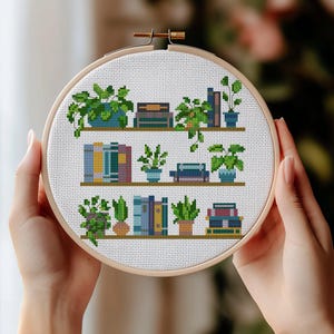 May include: A cross-stitch embroidery hoop depicting bookshelves with potted plants and stacks of books. The books have colourful spines, and the plants are green. The embroidery is set in a wooden hoop.