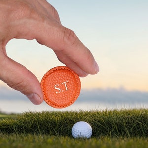 Può includere: Un segnapallina da golf rotondo in pelle arancione testurizzata con le iniziali bianche S.T. in rilievo al centro. Il segnapallina presenta un bordo cucito ed è tenuto sopra una pallina da golf bianca appoggiata sull'erba verde.