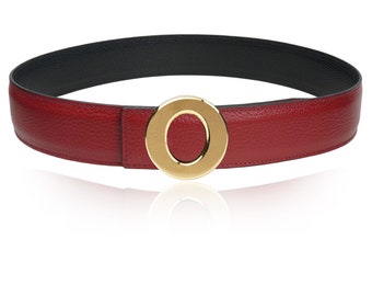 Puede incluir: Un cinturón de cuero rojo con una hebilla circular dorada. El cinturón es reversible con un lado de cuero negro.