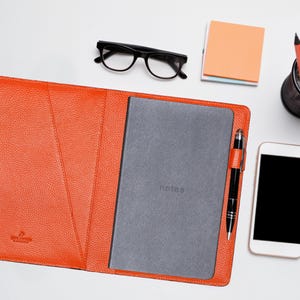 May include: An open orange leather notebook cover with a gray notebook and a black pen. A smartphone, glasses, sticky notes, and a pencil holder with pencils are also on the white surface.