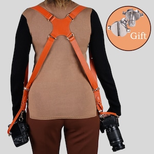 May include: An orange leather dual camera harness with adjustable straps and metal hardware, worn over the shoulders to support two cameras. A circular inset shows two silver metal mounting screws labeled with the word Gift.