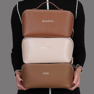 Puede incluir: Tres neceseres rectangulares apilados. La bolsa superior es marrón con el nombre "JENNIHER" en plata. La bolsa del medio es de color crema con "MICHAEL" en plata. La bolsa inferior es marrón con "JOHN" en plata.