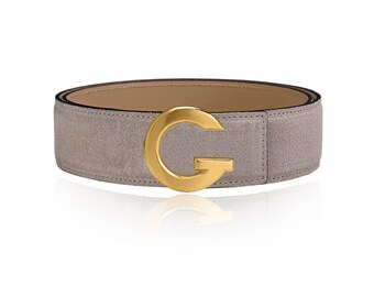May include: A light gray suede belt with a gold-colored metal buckle in the shape of the letter "G". The belt has a neutral-colored interior and a black edge. The buckle is centered on the belt.