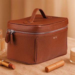 Puede incluir: Un neceser de cuero marrón con asa, cremallera plateada y las iniciales "MW" grabadas en la parte delantera. Un frasco de perfume, un labial y un tarro de crema están al lado.