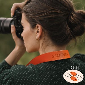 Può includere: Una tracolla per fotocamera in pelle arancione brillante con il testo SEMENTA in rilievo e cuciture visibili lungo i bordi. Un riquadro grafico mostra due piccoli attacchi triangolari in pelle arancione abbinati con passanti neri, etichettati come Gift.