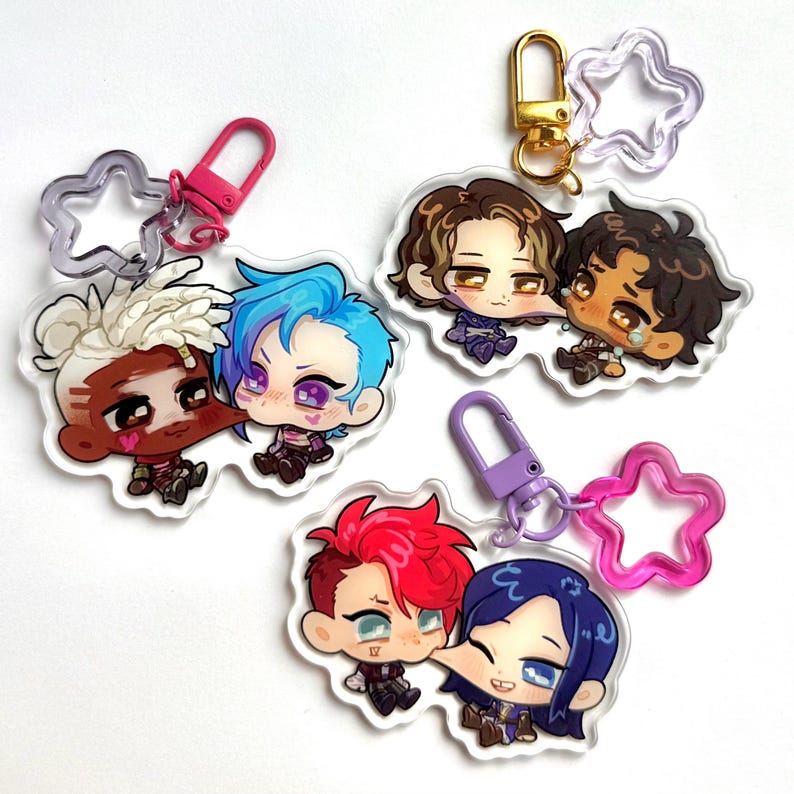 May include: Five acrylic charm keyrings featuring cartoon characters with blue, red, and brown hair. Each charm has a different character design and is attached to a metal clasp and a star-shaped charm.