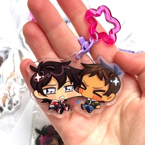 May include: A clear acrylic charm featuring two cartoon characters with black and brown hair. The charm has a pink star-shaped clasp and a purple clip. The characters are in chibi style, with one wearing a red and black outfit and the other in a blue jacket.