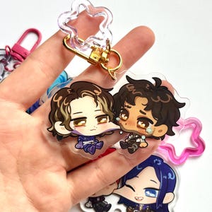 May include: Clear acrylic keyrings featuring cartoon characters with gold-coloured hardware. The keyrings have a star-shaped top and a gold clasp. The characters have detailed facial expressions and are wearing various outfits.