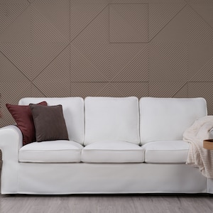 Op de afbeelding: Een witte bank met drie kussens en twee sierkussens, een bordeauxrood en een bruin, in een woonkamer met een textuurwand, een vaas met gedroogd pampasgras, een rieten mand, een lantaarn en een bijzettafel met een deken, decoratieve kaarsen en een gouden metalen sculptuur.