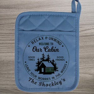 May include: Blue quilted pot holder with a white circle design. The circle has black text that reads "Relax & Unwind, Welcome to Our Cabin, Good Times, Good Friends, Leave Your Worries in the Woods, The Shockley's". Inside the circle is a black silhouette of a cabin with trees.