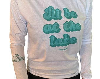 May include: White long-sleeve t-shirt with a teal green graphic that says "I'll be at the lake" and "Lake Surfer" in smaller text below. The left sleeve has a teal green wave graphic.