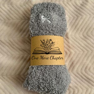 May include: A pair of gray fuzzy socks with a white bow detail. The socks are wrapped with a brown paper band featuring an open book illustration and the text "One More Chapter". The socks are on a light pink textured surface.