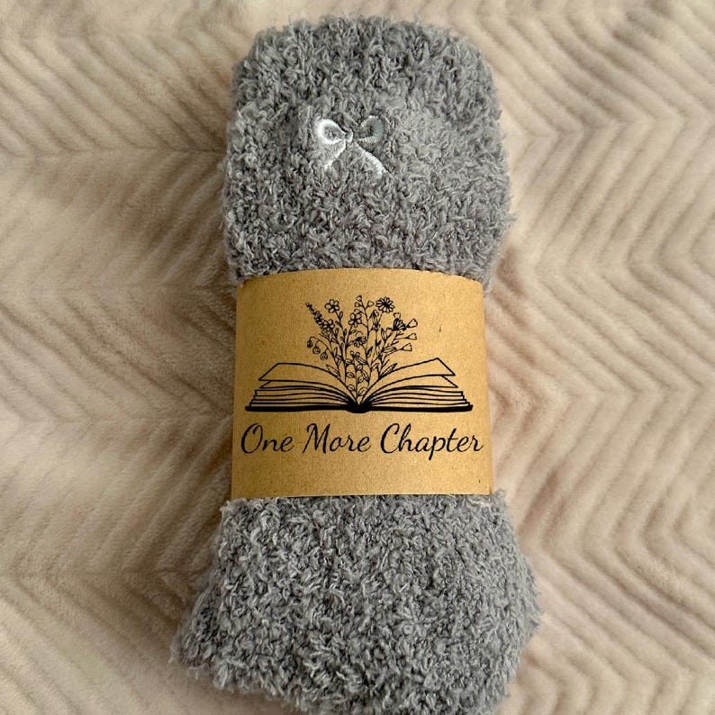 May include: A pair of gray fuzzy socks with a white bow detail. The socks are wrapped with a brown paper band featuring an open book illustration and the text "One More Chapter". The socks are on a light pink textured surface.