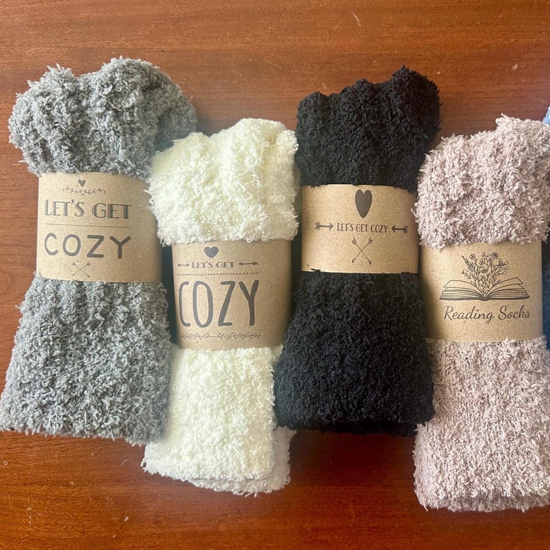 Pu&ograve; includere: Quattro paia di calzini pelosi con etichette di carta marrone. Le etichette hanno il testo "Let's Get Cozy" e un design a forma di cuore. Un paio di calzini &egrave; grigio, uno &egrave; bianco, uno &egrave; nero e uno &egrave; marrone.