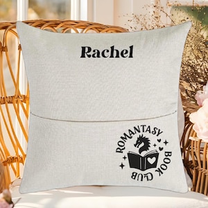 May include: A white pillow with a pocket on the back. The pillow has black text that says "Rachel" and a black graphic of a book, dragon, and the words "Romantasy Book Club".